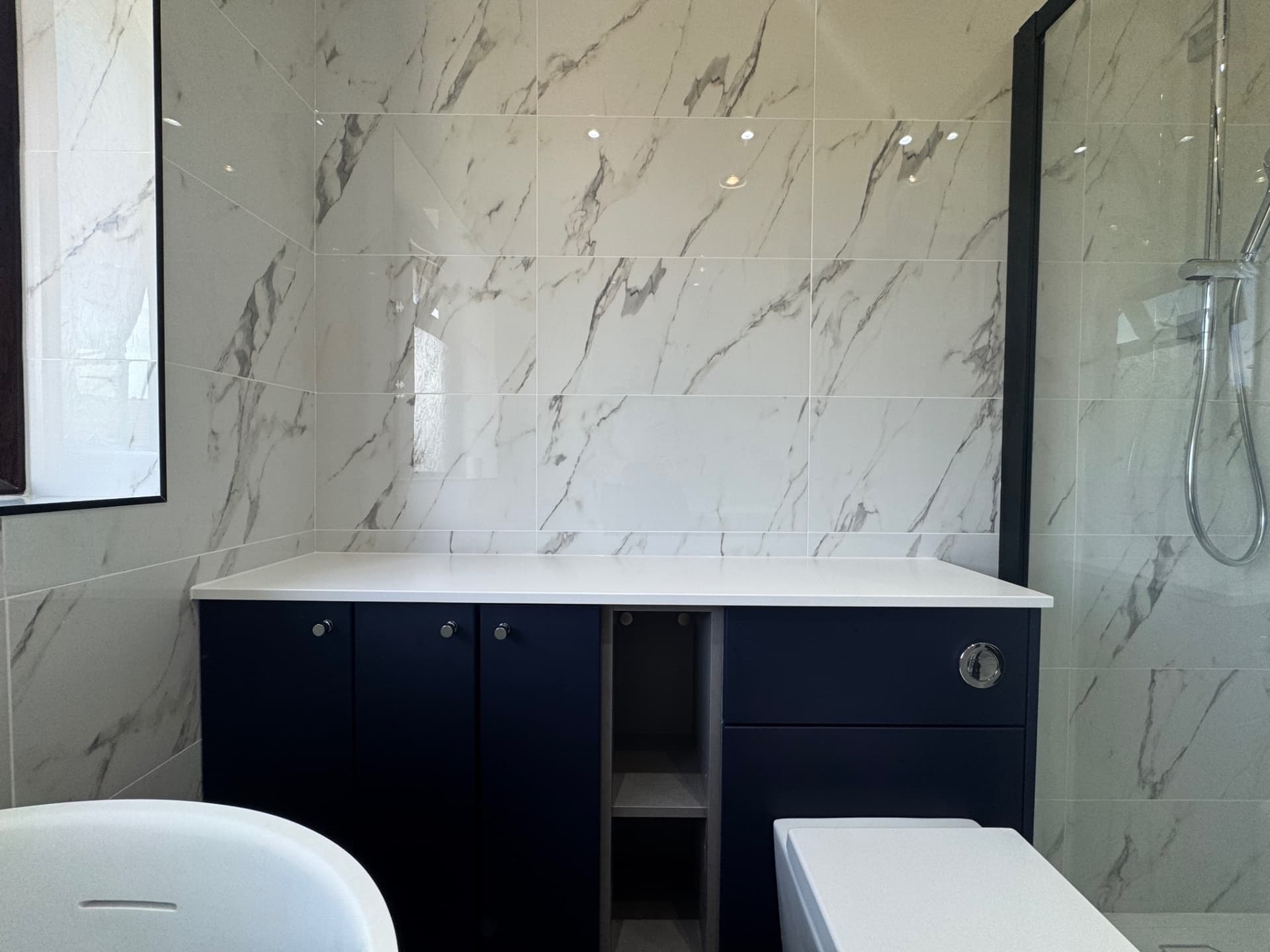 We've just finished installing this elegant bathroom with marble-effect tiling, a sleek navy vanity unit. The marble-effect wall creates a real focal point, while the contrasting dark cabinetry and white countertop give it a contemporary feel.